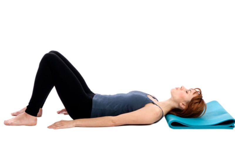 pilates boost digestion - A female wearing gym clothes lying on her back with arms by her side and knees raised. She has her eyes closed and her head is resting on a folded blue floor mat. On a white background.