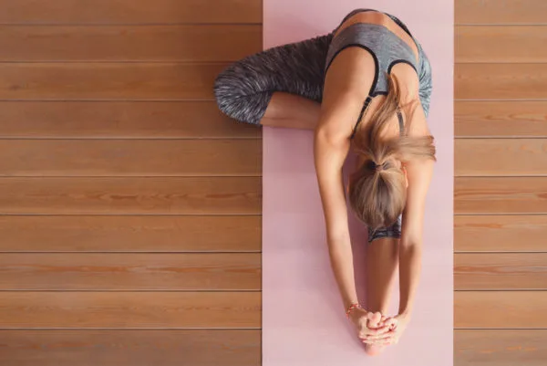 Exercise and cancer - An overhead shot of a woman wearing grey gym clothes performing a leg stretch on a pink gym mat on wooden flooring