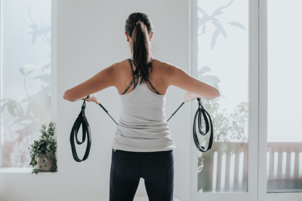 Surprising Benefits Of Pilates - A female in gym gear pulling straps with arms bent at the elbows facing away from the camera in a white washed studio with full length windows