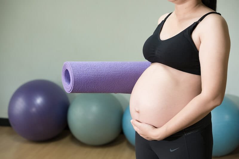 Pregnancy Pilates near me – Complete Pilates Prenatal Pilates London - a woman performing pilates exercises on studio floor with her feet and legs up on a silver gym ball in front of a white washed wall of full length net curtains and large window frames with the sunlight streaming in.