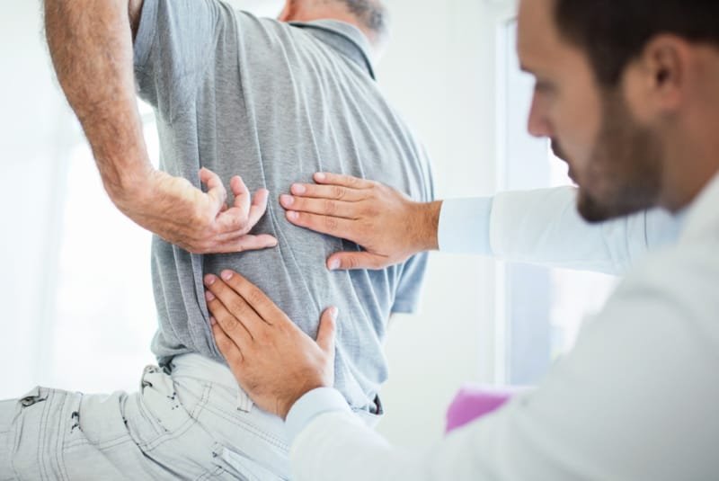 Back Pain - A man with back pain sitting on a bench in a doctors office with the doctor placing his hands on the painful area.