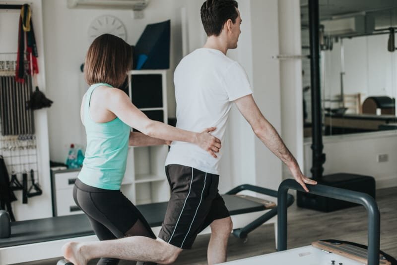 Benefits of Clinical Pilates - A make and female wearing gym clothes in a Pilates studio. The male is being assisted by the female as she has both her hands holding his hips. He has one foot on a Pilates reformer machine facing away from the female and using one hand holding a bar to balance.