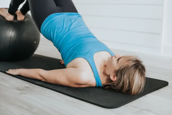 Pilates bridge on a ball - A female wearing gym clothes lying on a floor mat with arms by her side and feet placed on a stability ball