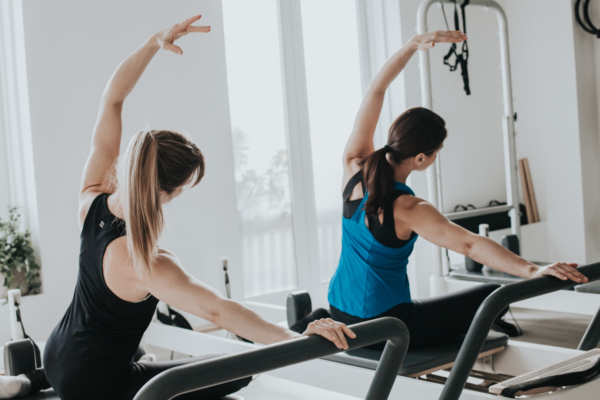pilates myths - two females facing away from the camera with their left arms arced over their heads while the right arms arm straight against a pilates bar and they lean towards the bar with their torsos