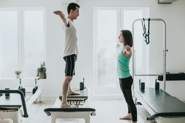 Surprising Benefits Of Pilates - a male and female practitioner of pilates in a pilates studio standing facing each other with arms and legs outstretched to the sides with the male standing on a bench