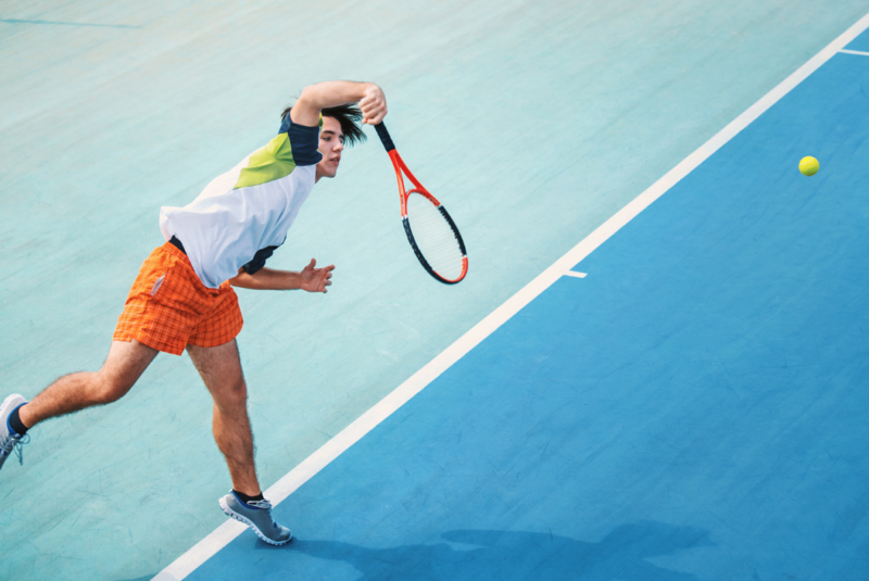 Pilates improves sports performance - An overhead shot of a clay tennis court coloured two shades of blue. A young man is in mid-swing having just hit the tennis ball which can be seen in shot to the right of the frame. The man is wearing orange shorts and a white t-shirt with green and blue on the sleeves.