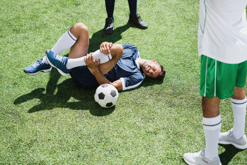 Pilates improves sports performance - An overhead shot of a footballer lying on the pitch holding his left leg and fake grimacing in pain as though he has really injured himself. A football rests beside him and there are two other footballers standing in attendance. You can only see their legs and feet. All are wearing different coloured shorts, tops and trainers.