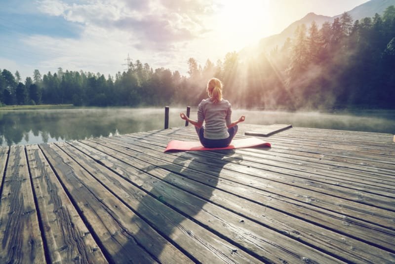 Setting intention - A female itting on a mat on a wooden pontoon overlooking a lake fringed with wooded slopes at sunrise. She is sitting in the Lotus position with her back to the camera.