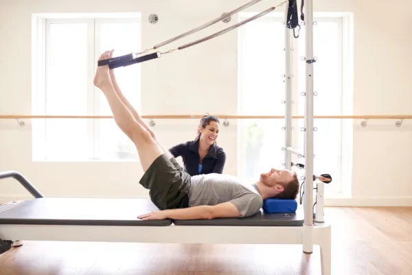 Trapeze table - A Pilates practitioner lying on his back on a trapeze table with his feet attached to straps held above him.
