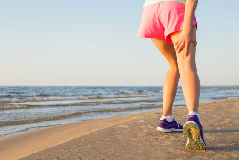 Tight hamstrings - A female holding her hamstring with her hand. She is on a sandy beach by the waterline in broad daylight, wearing pink shorts and dark purple trainers.