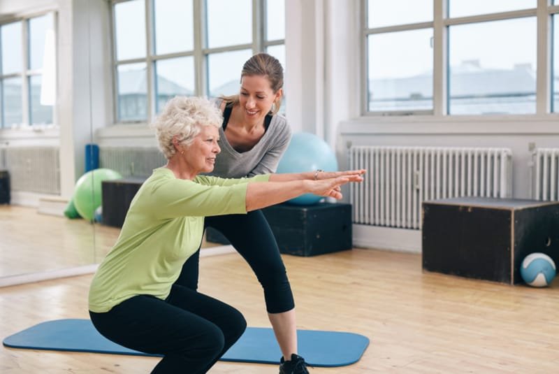 Why women need strength training - An older female performing squats in a studio being guided by a younger female