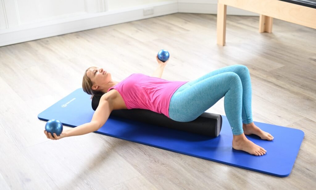 Woman lying on foam roller, using Pilates weights to stretch her chest and shoulders